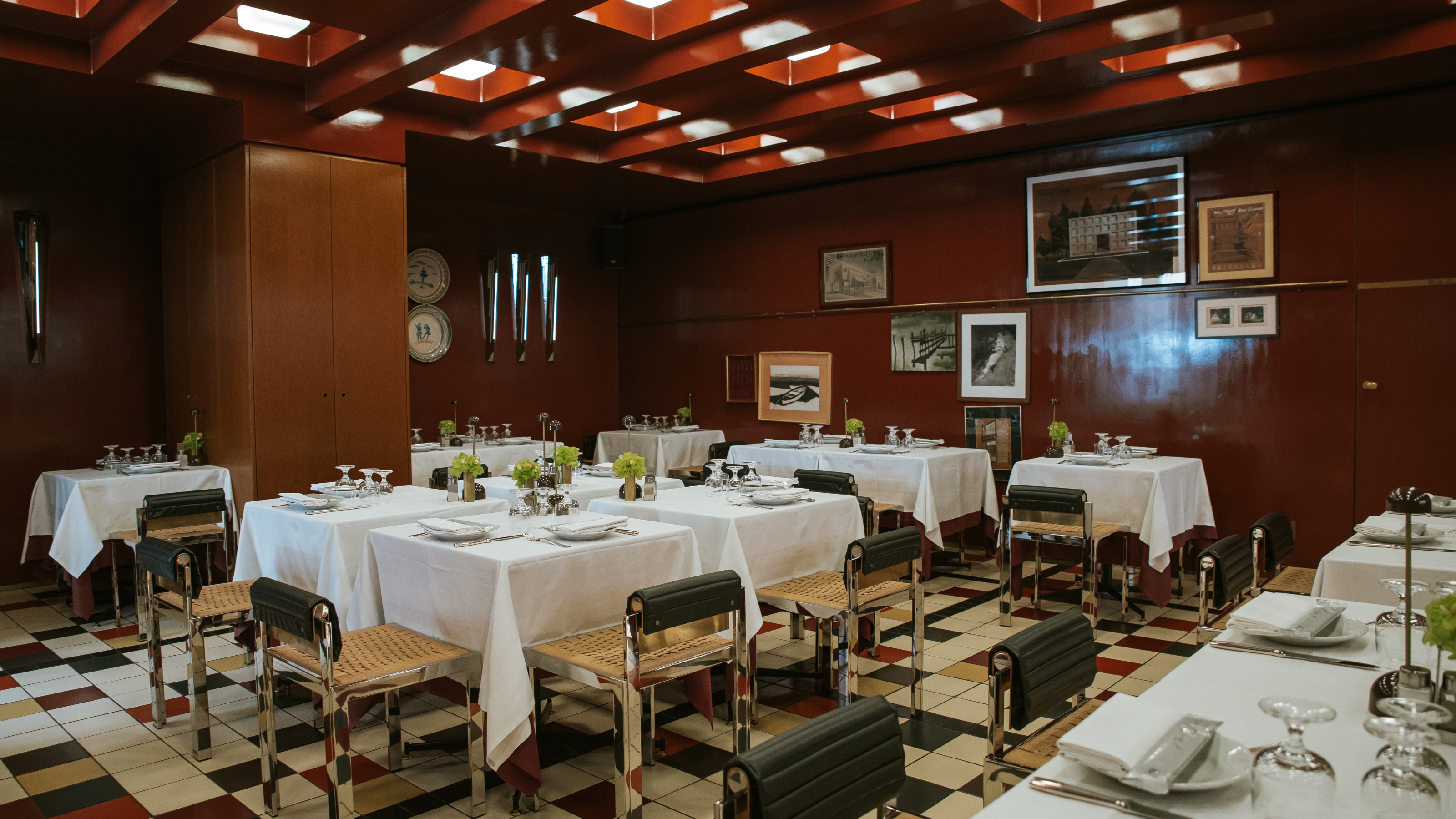 A 1960s-inspired restaurant with coffered, back-lit ceilings, a terracotta-hued palette, checkered floors in red, black, beige, and white, white clothed tables, and rattan, chrome, and leather chairs.