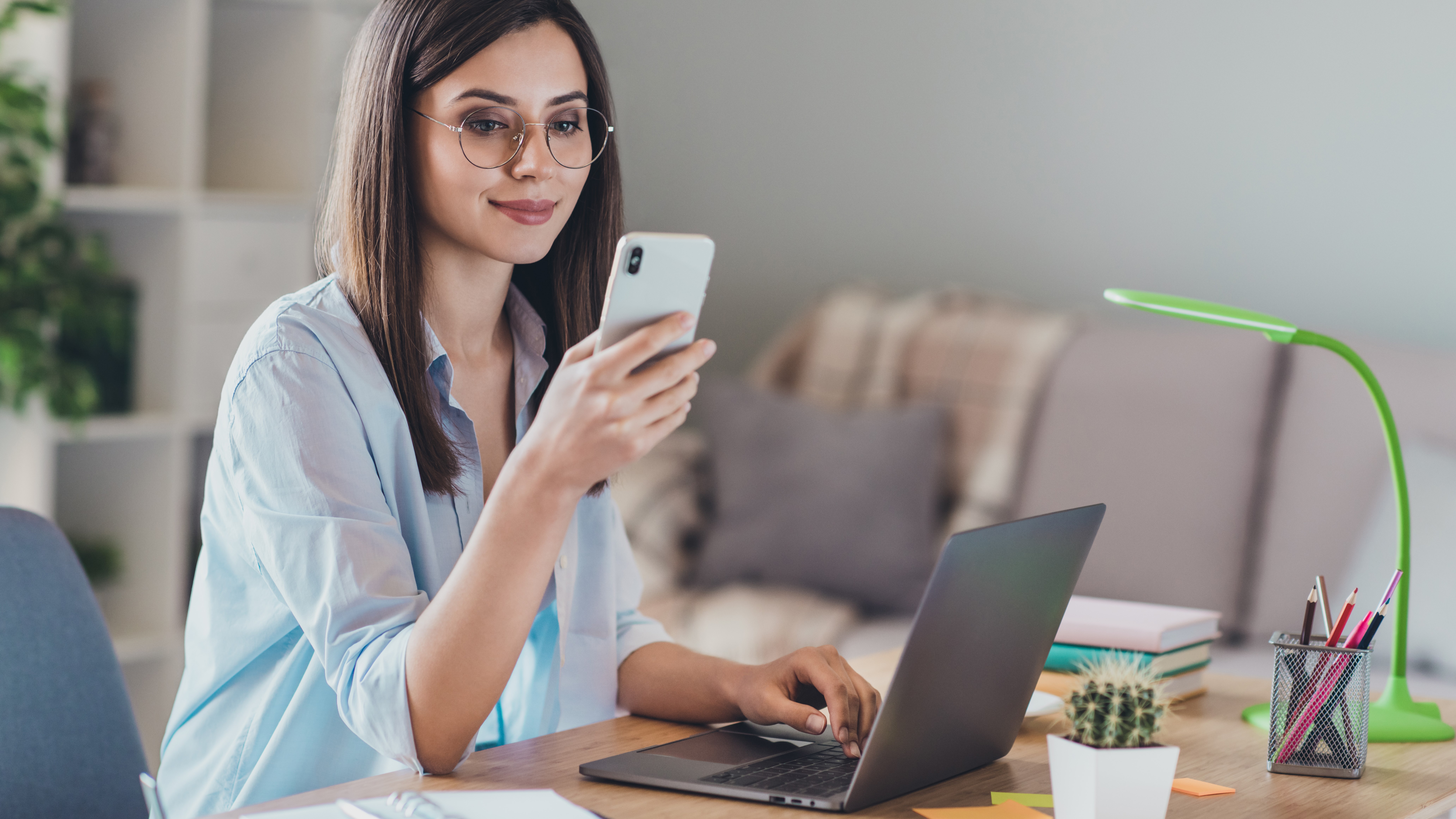 Mujer sentada en un escritorio con una computadora portátil, sosteniendo un teléfono y mirándolo