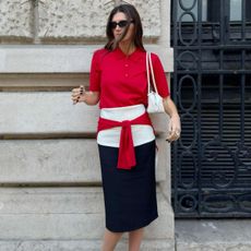 Woman wears red polo top, white belt, black skirt and carries white bag