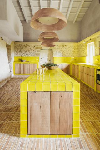 rustic style yellow kitchen with stone wall, brick floors, timber cabinetry, and neon yellow tiled kitchen counter and backsplash. timber dome-shaped pendant lights hang over the kitchen island