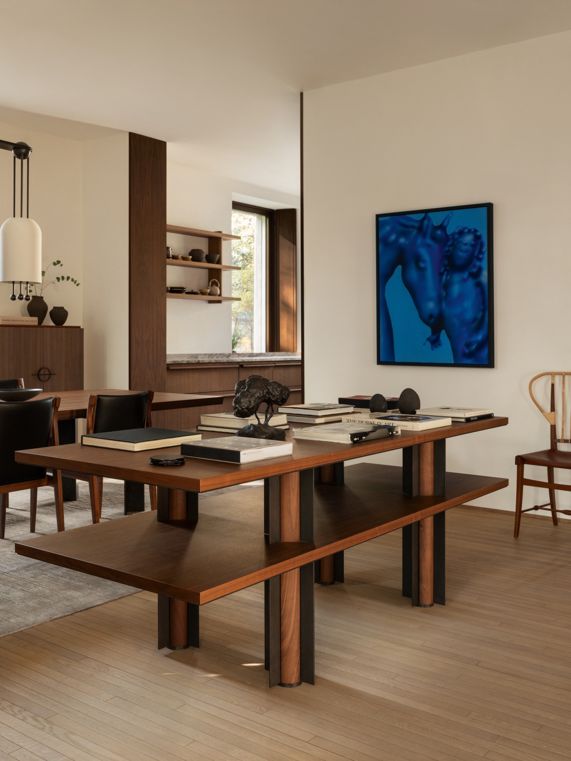 Two-tier wooden library table in a room with wooden dining table and chairs and figurative cobalt blue artwork