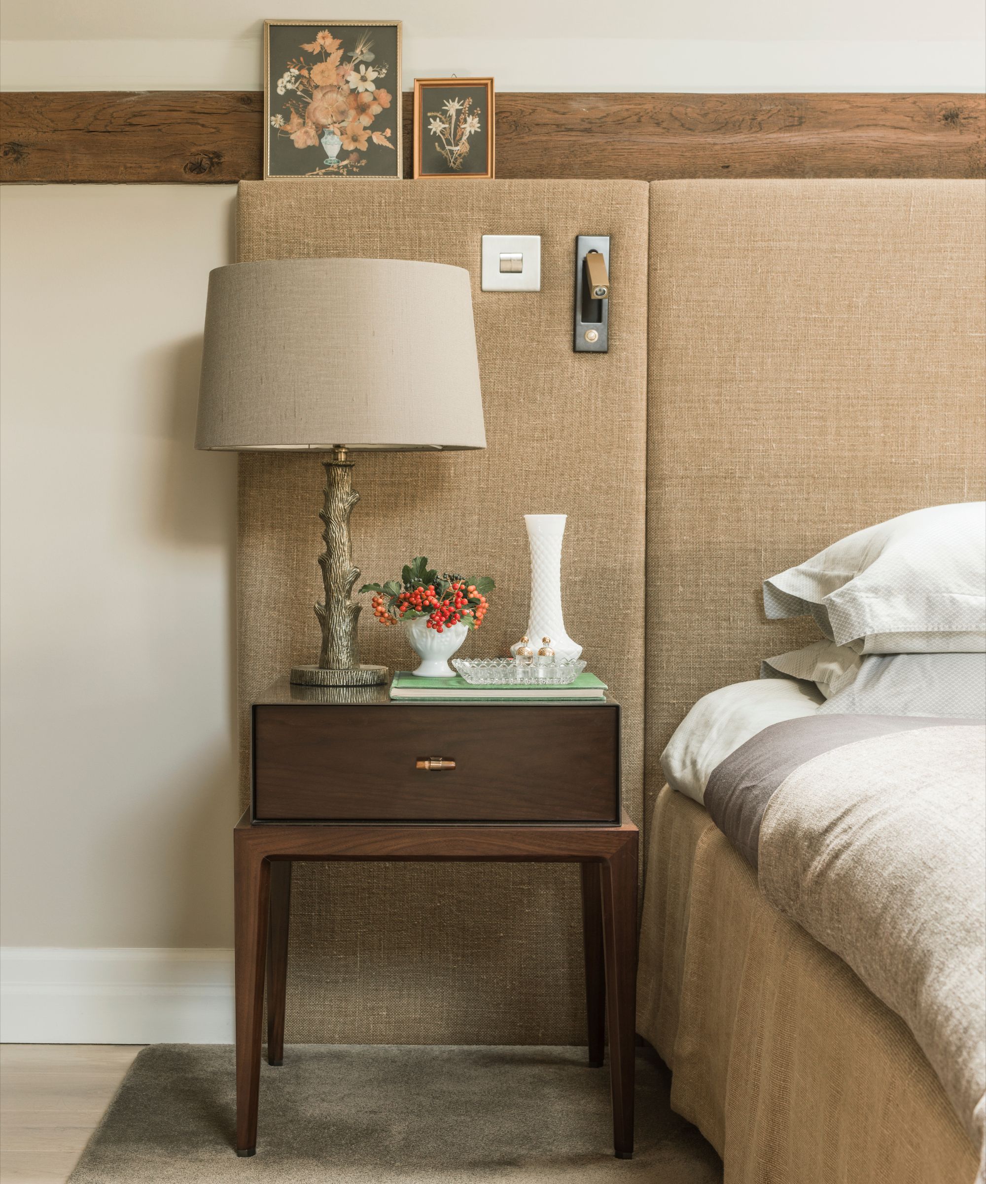 A neutral bedroom with a grey rug on wooden flooring, a brown dark wood bedside table with glowers, a vase, and a lamp, with white bedding and a padded headboard.