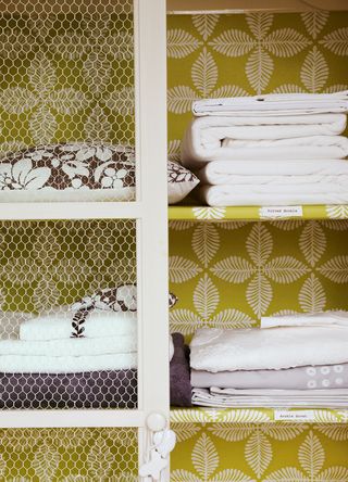 Picture of a laundry cupbaord with white sheets and pillows on shelves with a green wallpapered background to support the Dunelm take Back scheme for recycling home textiles