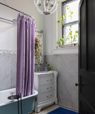 A bathroom with gray marble on the lower half of the walls and the upper half painted warm white. A white vanity sits in the far corner, while freestanding light blue bathtub is places next to it with a purple shower curtain suspended above from a silver ceiling mounted rail