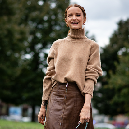 A guest attends milan fashion week spring summer 2025 in beige turtleneck sweater and brown leather skirt smiling 