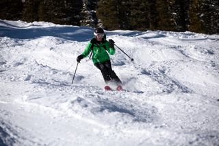 A woman in a green coat and black pants skiing down a run with moguls