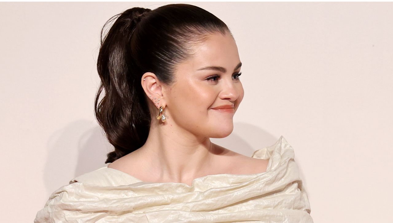 Selena Gomez wearing a wrinkled off the shoulder white dress in front of a plain backdrop