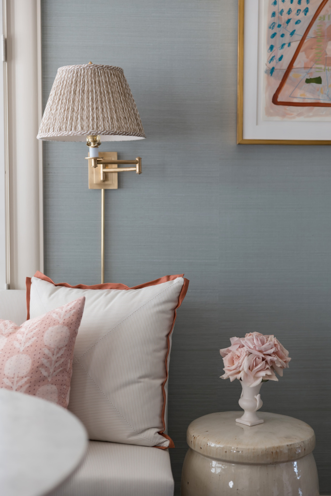 A wall sconce with a tan pleated lampshade sits above a white coach with pillows and a tan side table with flowers.