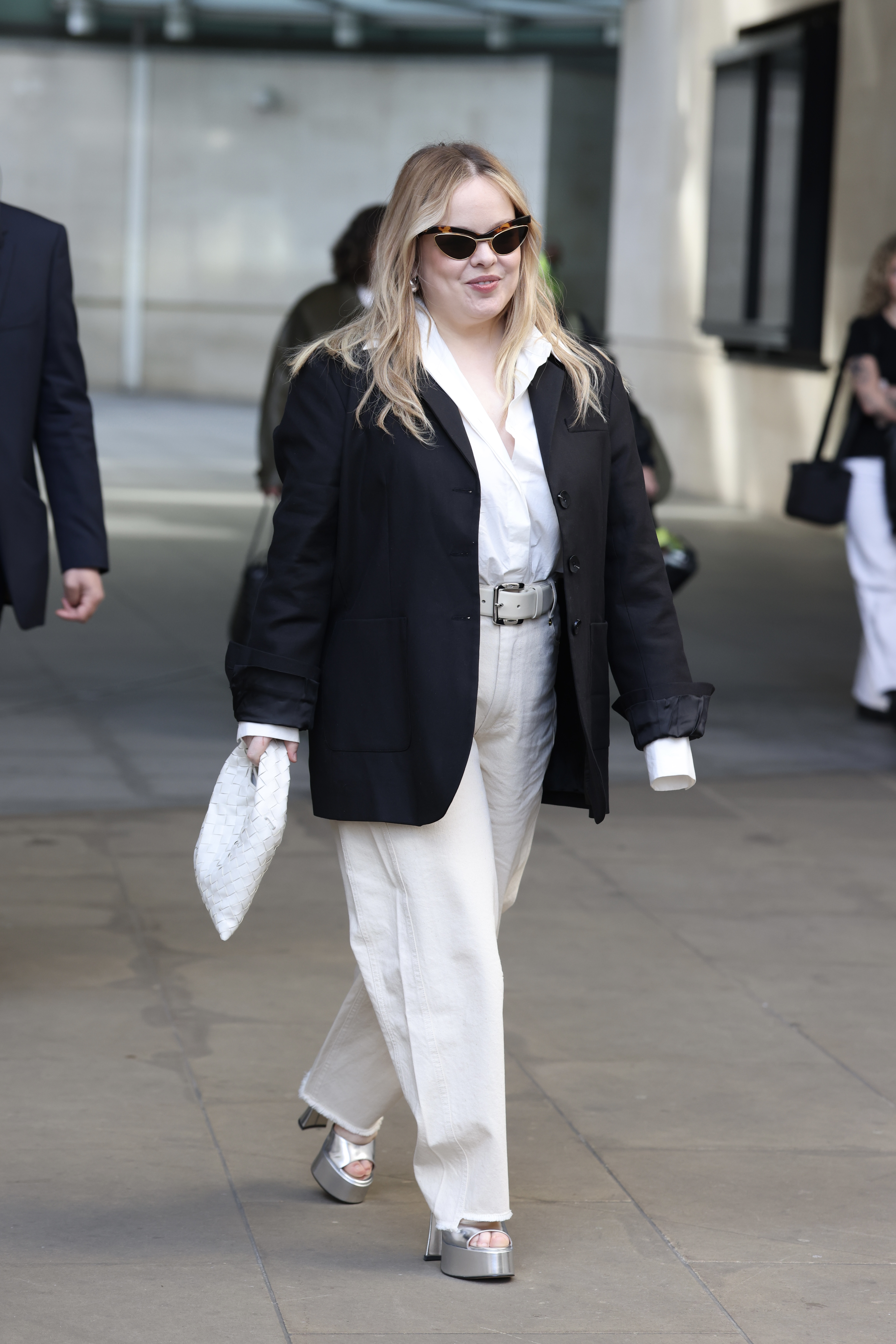 Nicola Coughlan walks down the street wearing white jeans with silver platform heels, a white shirt and a black blazer.
