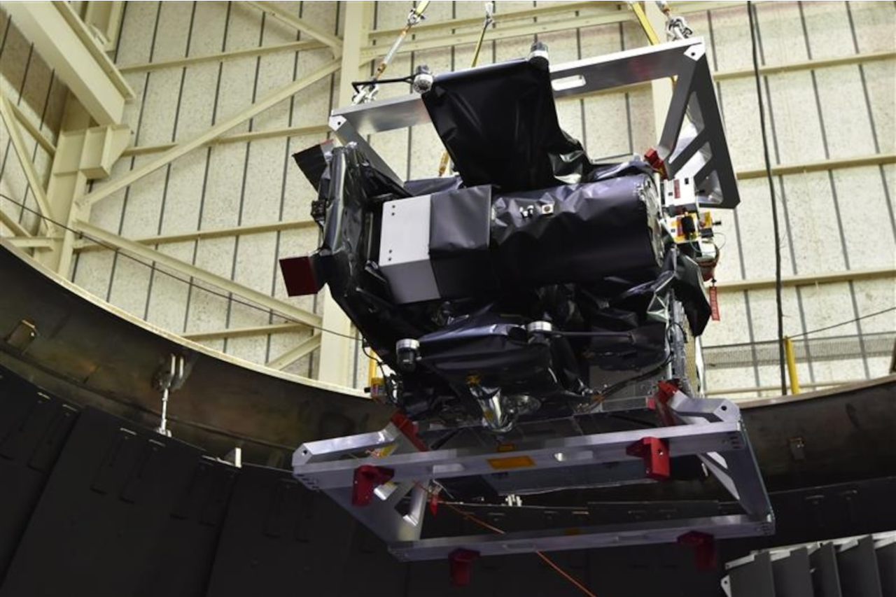 a roughly cube-shaped metal object consisting of a top and bottom square frame with electronic components in between. the object hangs from metal cables inside a hangar