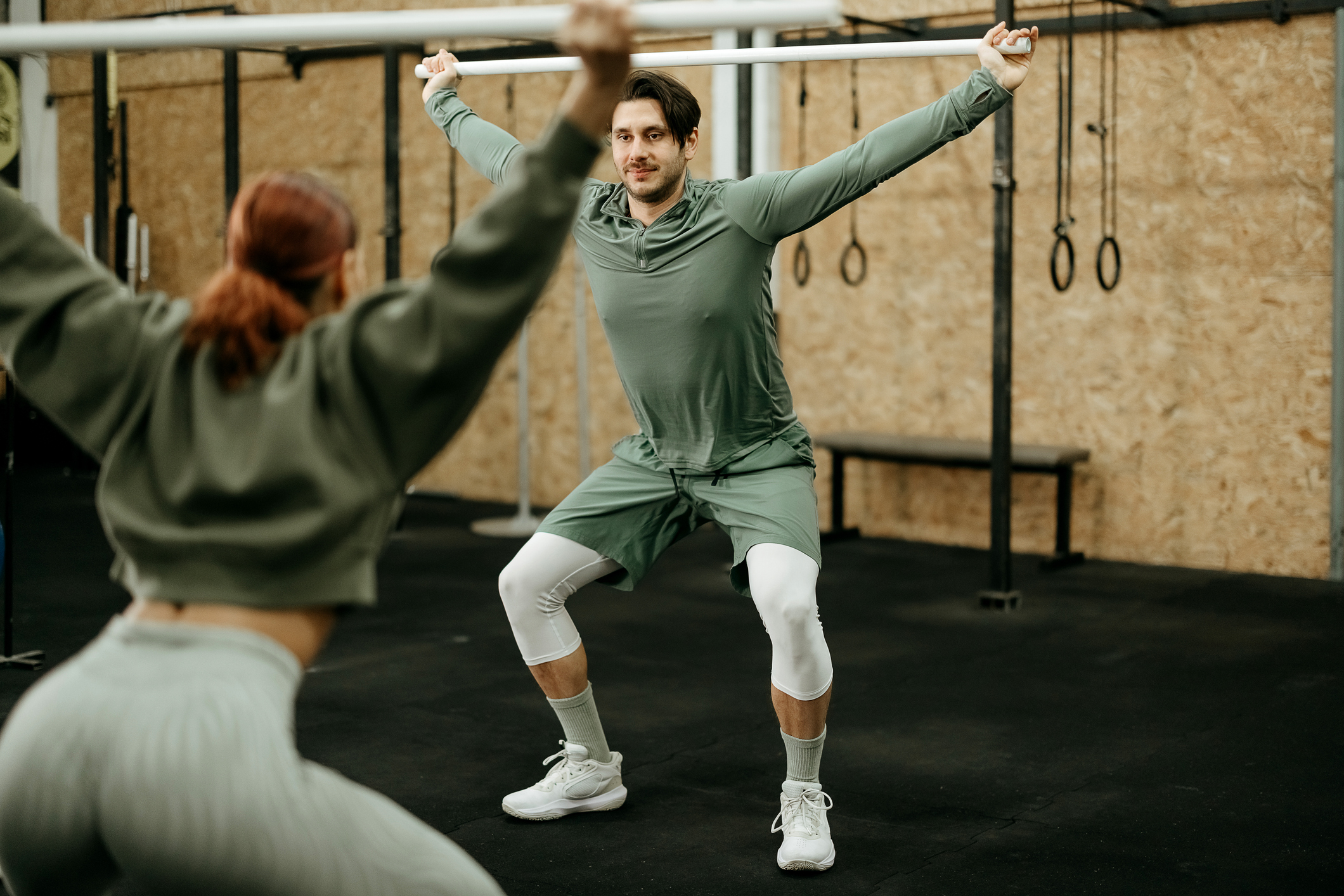 Two people exercising in a gym holding a plastic pipe overhead