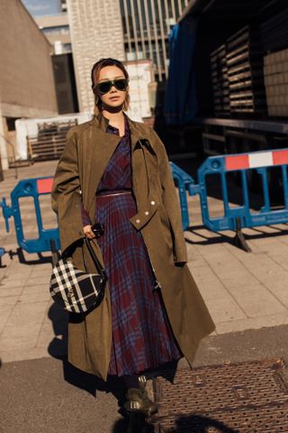 Fashion week guest wearing the English country style trend of a tartan dress, plaid, bag, and suede trench coat