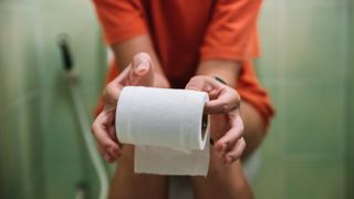 The mid-section of a person is shown as they are sat on the toilet holding a roll of toilet paper. 