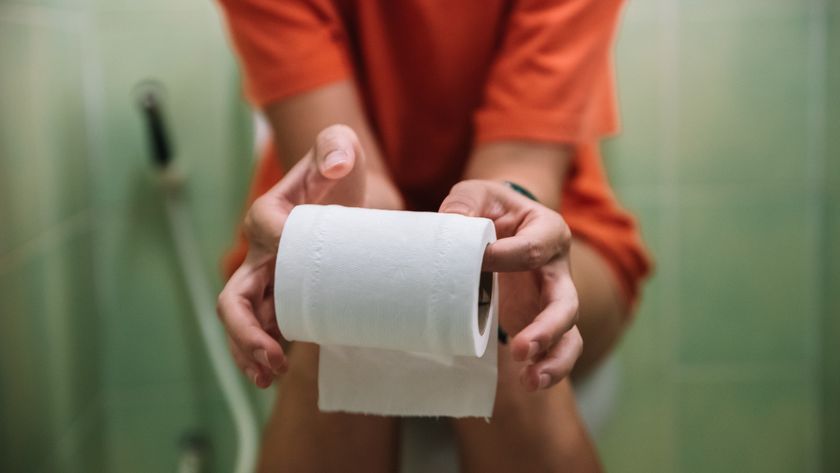 The mid-section of a person is shown as they are sat on the toilet holding a roll of toilet paper. 