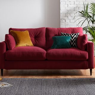 Burgundy red sofa with velvet cushions in front of a white brick while and on a black and white patterned rug