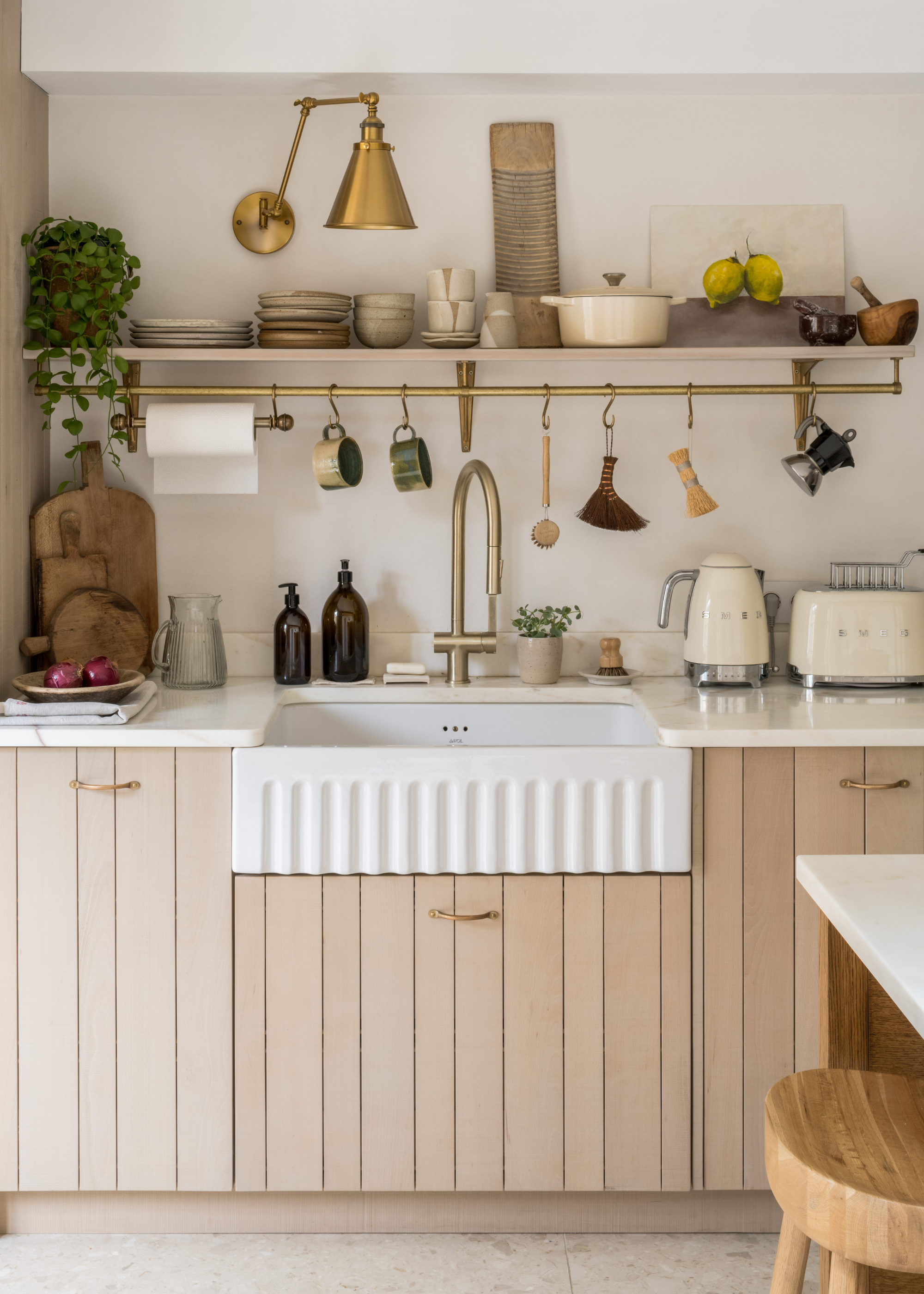 A kitchen with wooden cabinets a shite sink and a countertop with a SMEG kettle and toaster, a set of wood cutting boards. Above a hanging rod display with brushes, mugs and a moka pot along with open shelves holding a trailing plant, plates, cups, a pot and a painting