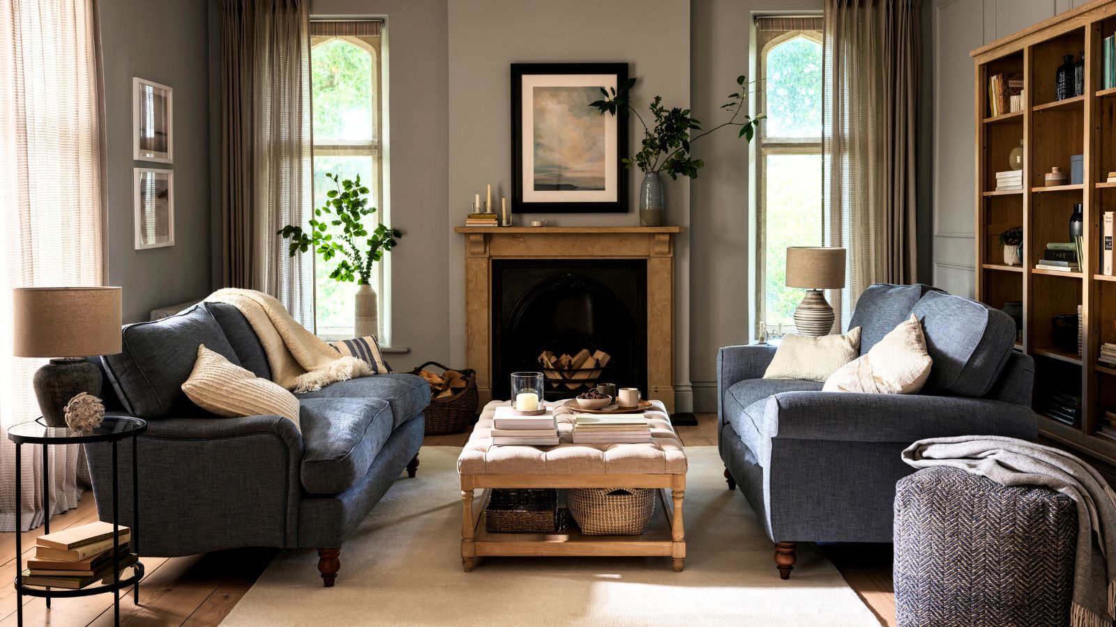 A cozy living room with two blue sofa&#039;s facing one another over an upholstered top coffee table. A wooden fireplace behind. 