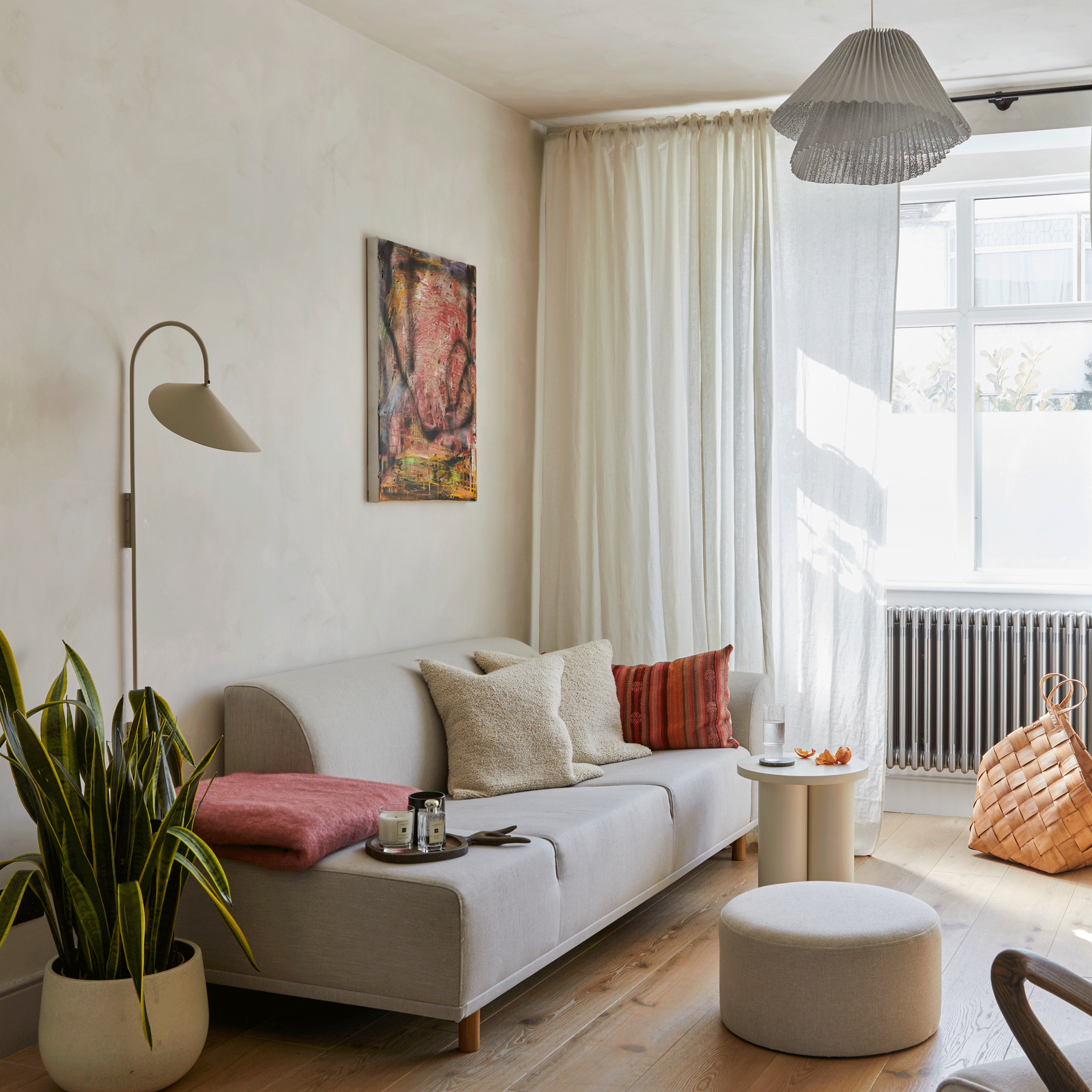 a living room with a plain modern sofa with read and cream cushions with a side table and foot stool and a abstract artwork on the wall