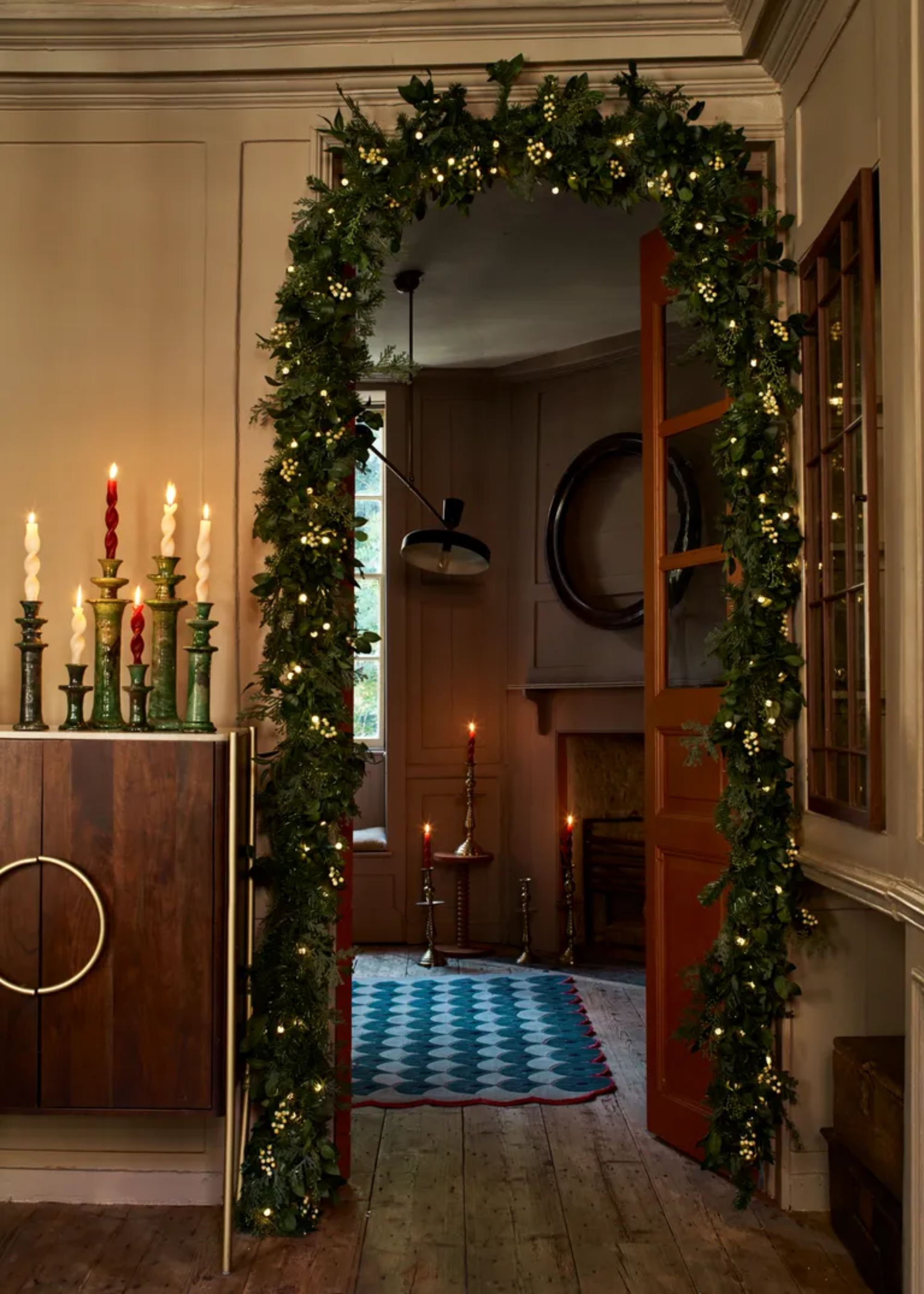 A doorway with an arched garland with baubles by a sideboard with lit twisted candles on ceramic tapers