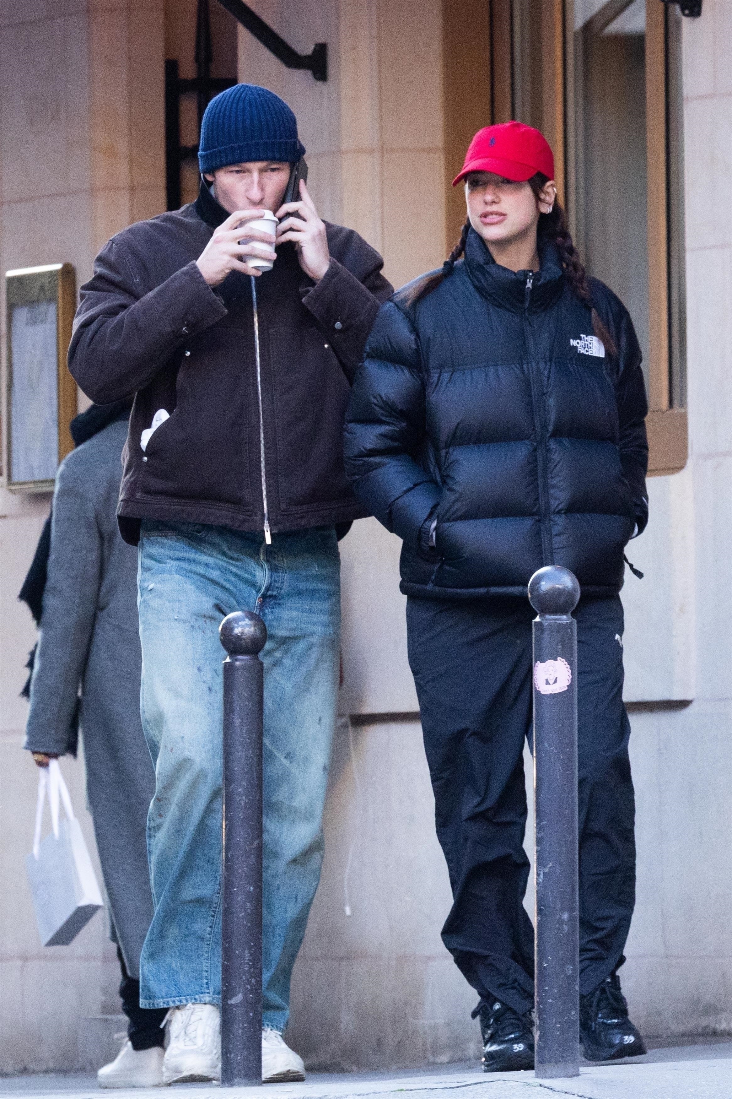 Dua Lipa wearing a The North Face black puffer jacket, black pants, black sneakers, and a red baseball cap