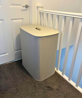 The Tota laundry organizer in my home, pictured on gray carpet in my upstairs hallway, with a white closed door behind to the left, and white painted staircase railing to the right