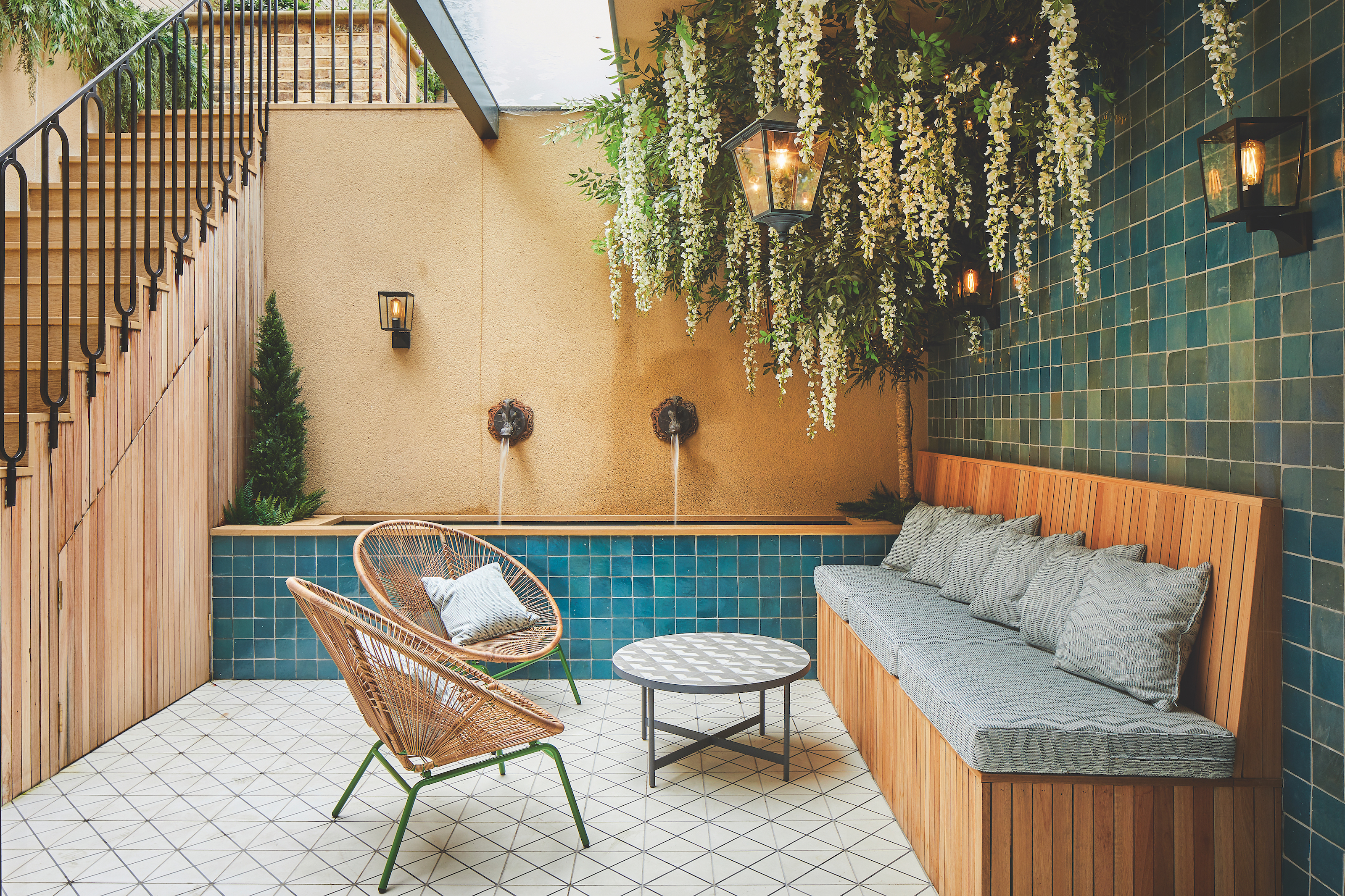 Courtyard area with blue tiles covering the wall and water feature, with a wooden bench seat below hanging wisteria. The floor tiles have a geometric pattern with two wicker chairs and a round coffee table in between.