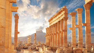 Ancient Roman time town in Palmyra, Syria. Mostly destroyed, we do see several columns standing tall. in the background there is a hill with a settlement on top against a blue, clouded sky.