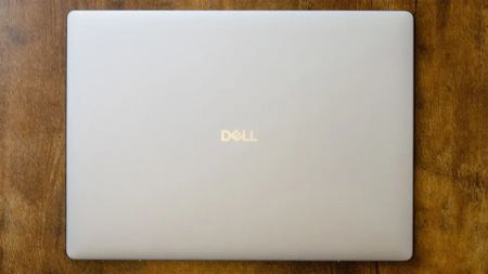 A closed silver Dell laptop rests centered on a warm, textured wooden table, viewed from above; the simple arrangement feels calm and uncluttered.