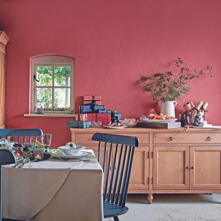 A red dining room decorated for Christmas. Sideboard is topped with presents, and the table is decorated with candles and greenery