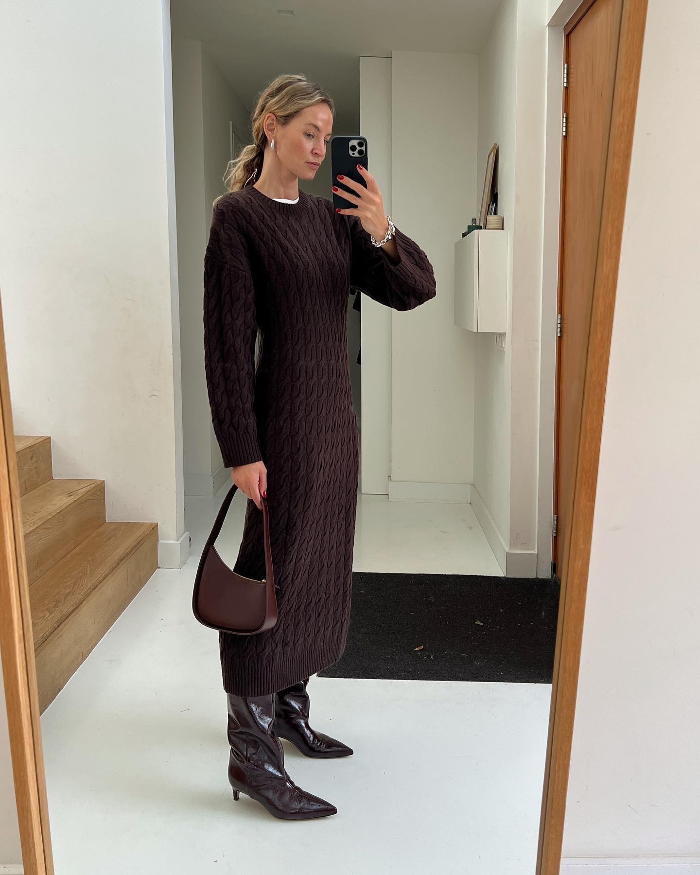 Anouk Yve is in a brown sweater dress, brown heeled boots, and a curved brown shoulder bag.