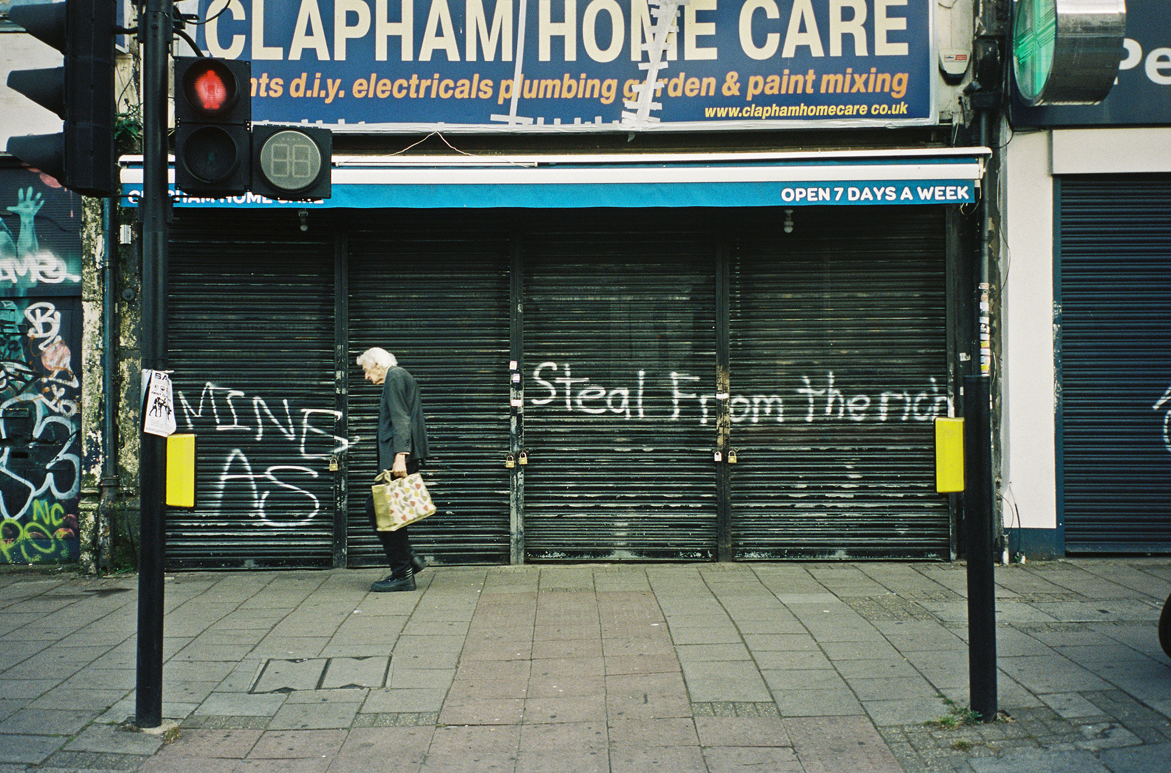 Colour photograph of an elderly person walking along the street past a shuttered shop front that has &amp;ldquo;STEAL FROM THE RICH&amp;rdquo; spray-painted in front