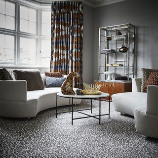 A pair of grey curved sofas arranged around a circular coffee table in a grey living room with animal print carpet.