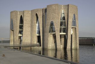 Fjordenhus, by Olafur Eliasson and Studio Olafur Eliasson in Vejle, Denmark. Modern face brick two storey building surrounded by water with triangular arched windows and a bridge.