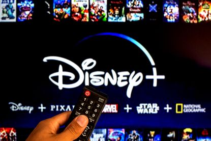 A close-up of a hand holding a TV remote control seen displayed in front of the Disney+ logo.