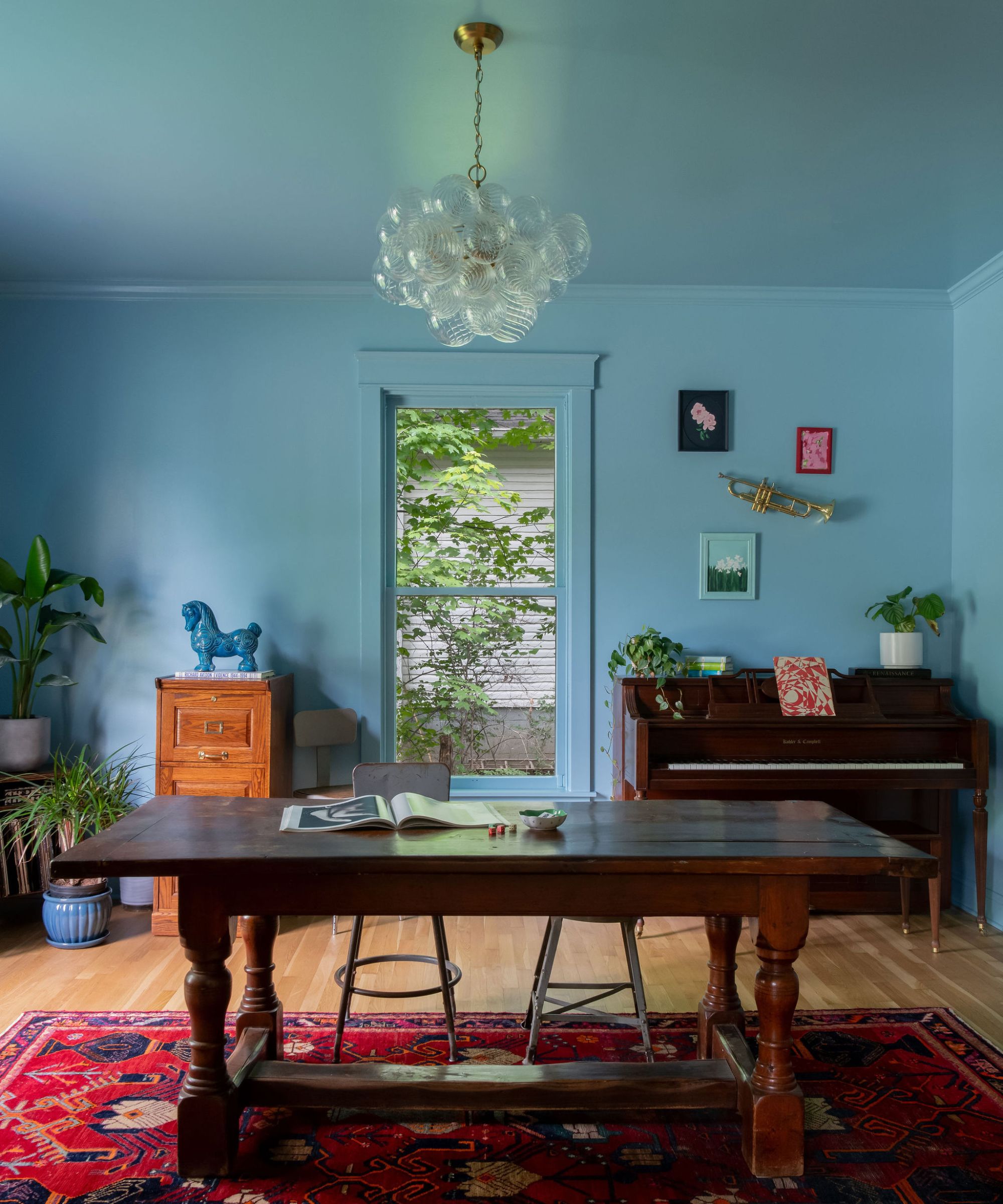A parlor painted blue with a large wooden desk, a red rug, and a piano in the corner.