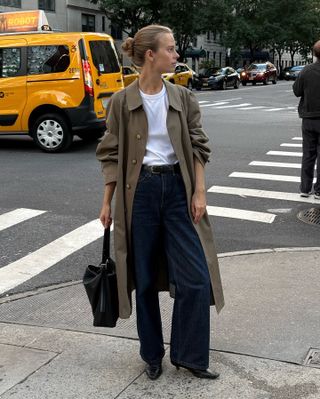 Scandinavian creative Clara Dyrhauge poses in NYC wearing an olive green trench coat, white t-shirt, a black belt, dark-wash jeans, and black boots.