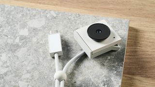 A photo of the Opal Tadpole tiny webcam in white and gray colorway, placed on a stone slate and wooden desk.