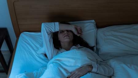 A woman lies in bed at night unable to sleep. She has her right arm over her face and her left arm resting on the duvet.