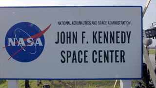 white sign that reads "john f. kennedy space center" in black all-caps type, with nasa's blue "meatball" logo to the left of the words