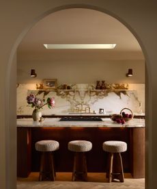 Arch doorway looking into modern kitchen with wood and marble round kitchen island with three round stools, matching marble backsplash with shelves and two wall lights