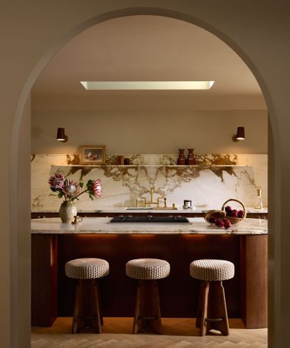 Arch doorway looking into modern kitchen with wood and marble round kitchen island with three round stools, matching marble backsplash with shelves and two wall lights