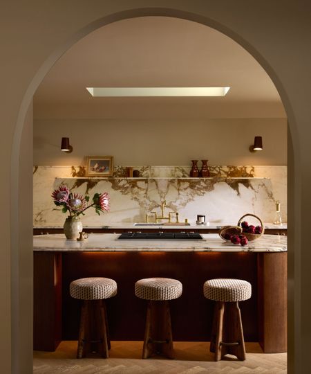 Arch doorway looking into modern kitchen with wood and marble round kitchen island with three round stools, matching marble backsplash with shelves and two wall lights