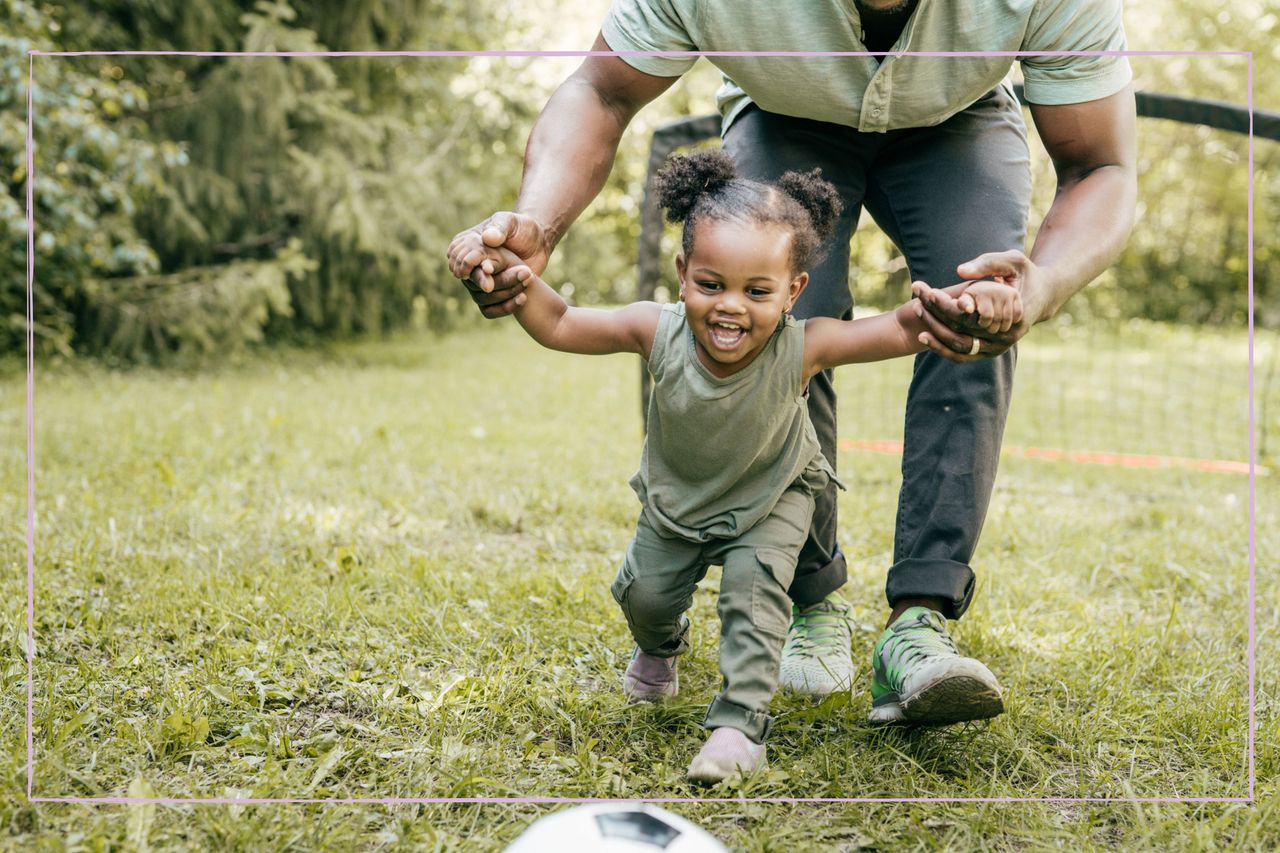 Happy toddler chasing after a ball in a garden, with her hands being held by her dad in support