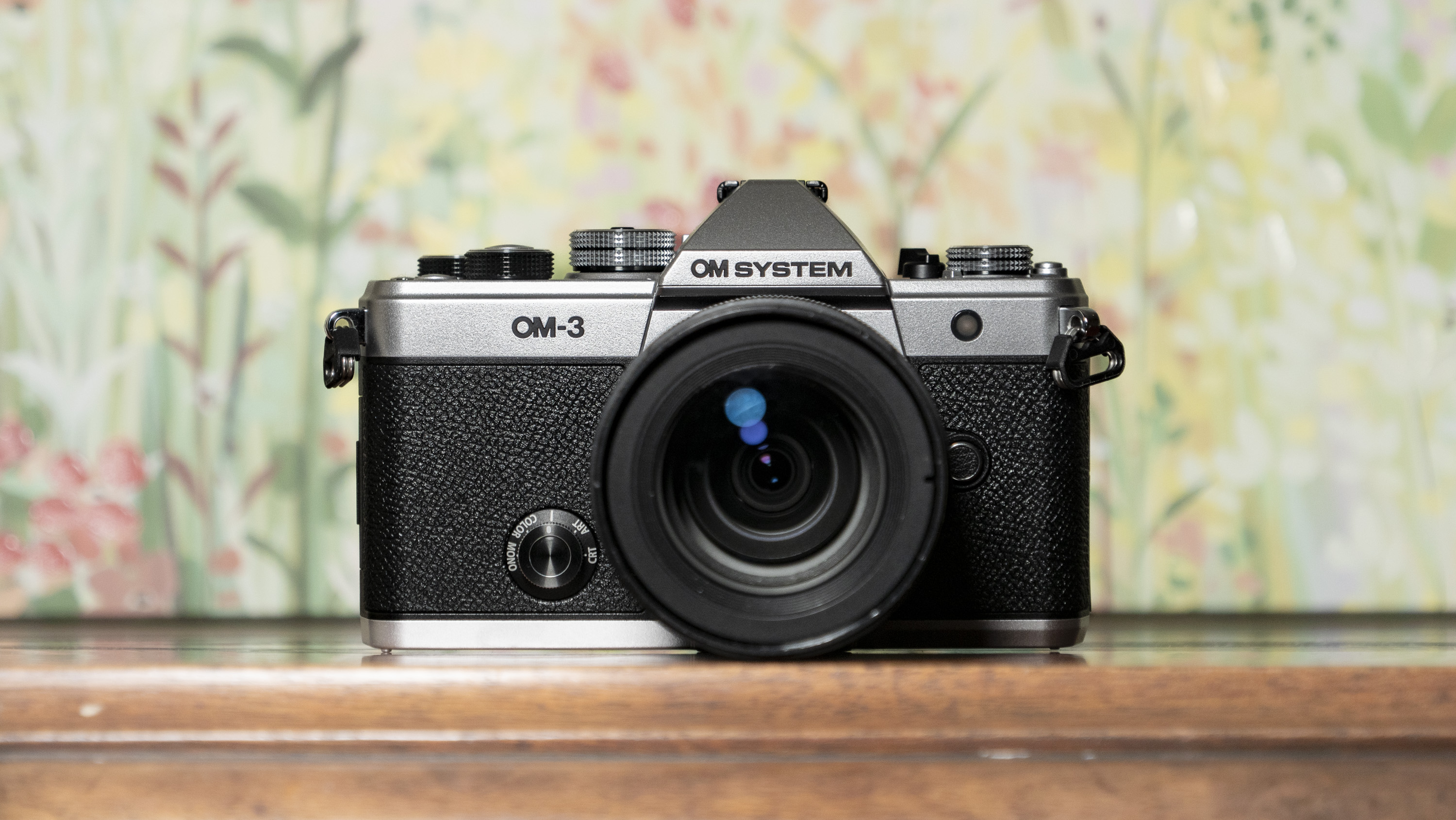 OM System OM-3 mirrorless camera on a wooden surface with floral painting background, 12-45mm F4 Pro lens attached