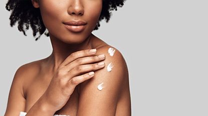 Close up of young woman applying body cream and smiling while standing against grey background