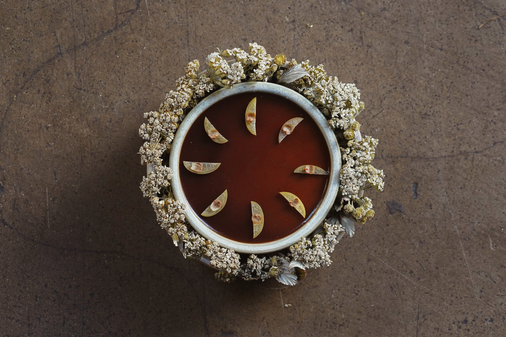 A sculptural dish filled with slices of fruit swimming in a brown dressing is decorated with natural blossoms and placed on a brown stone surface.