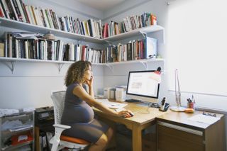 Woman looking at her laptop and on the phone in home office, researching the effects of the covid vaccine and pregnancy
