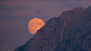 a large supermoon is partially hidden by a rocky mountain, the moon appears large and slightly orange.