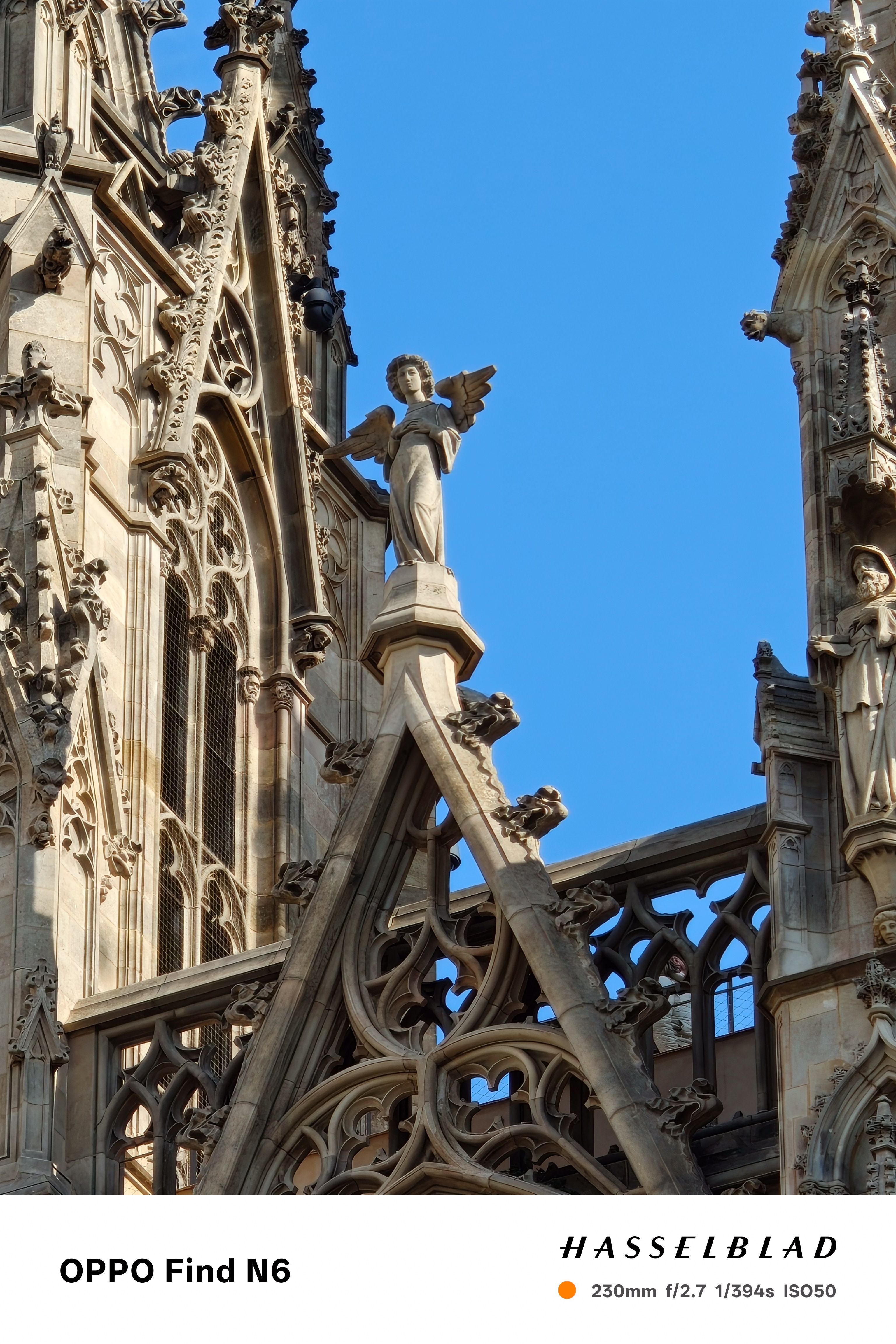 A medium-long shot of a weathered stone angel sculpture perched atop a Gothic-style gable. The angel is depicted with folded hands and large wings against a clear, deep blue sky. The intricate stonework and arches of the cathedral are visible in the background.