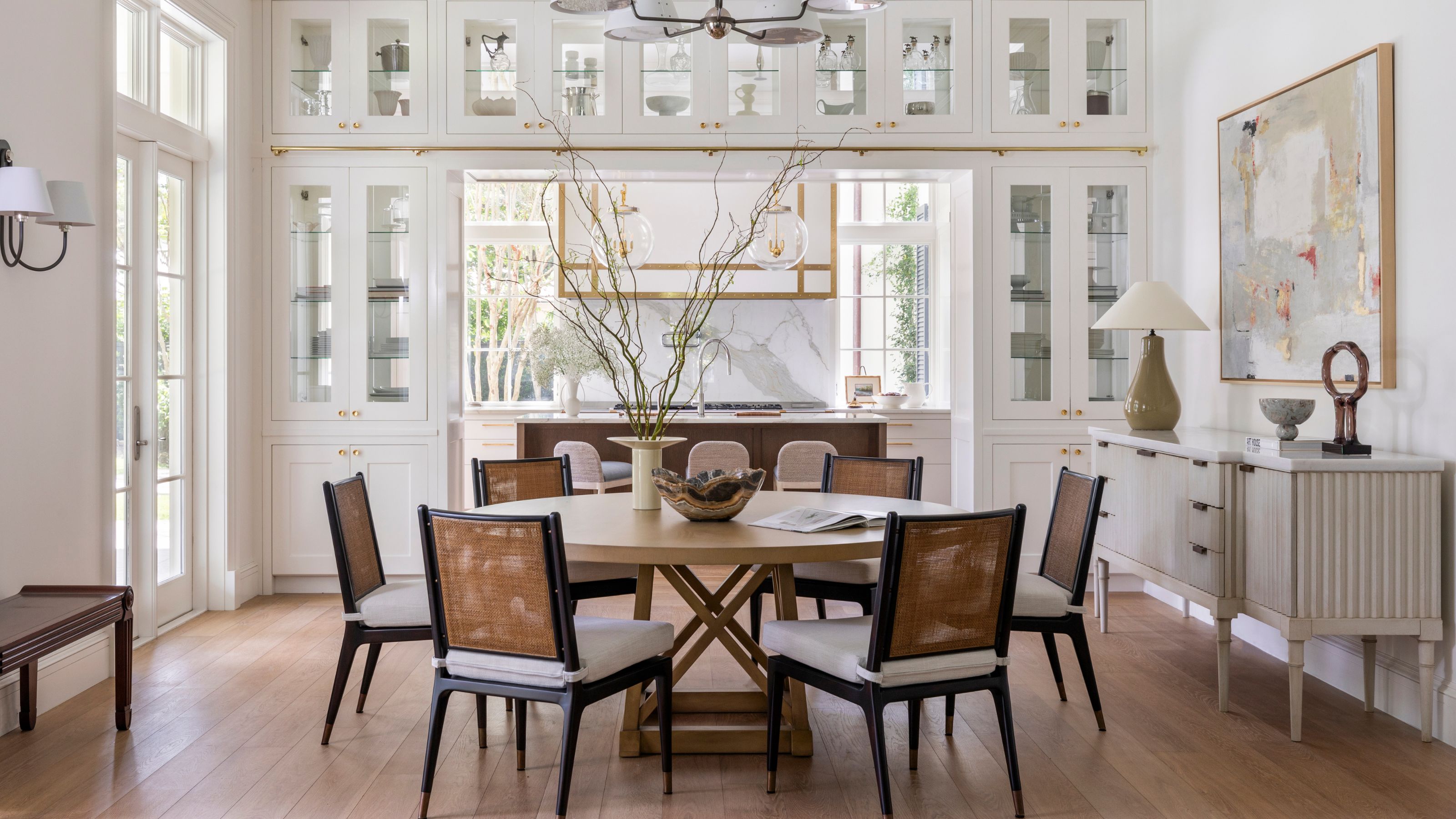 a large white and light breakfast area off of a kitchen with a walk through bookcase, round table and black and rattan chairs